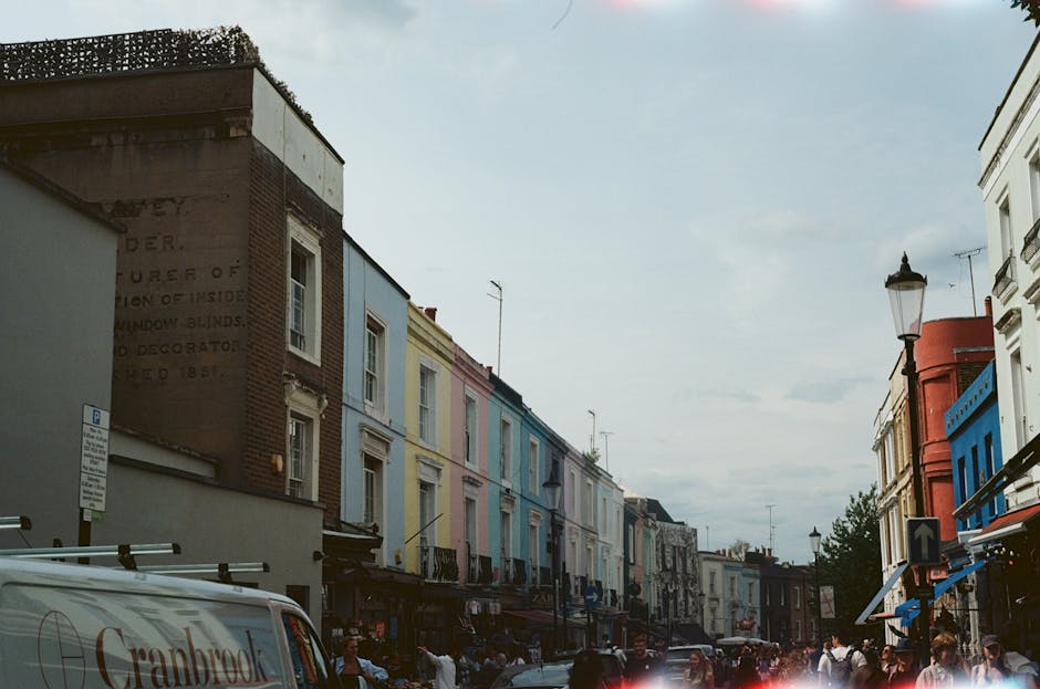 The image depicts a busy street scene in Notting Hill during daytime, with a row of parked cars along the curb on the left side and people walking along the sidewalk, some carrying shopping bags. The 