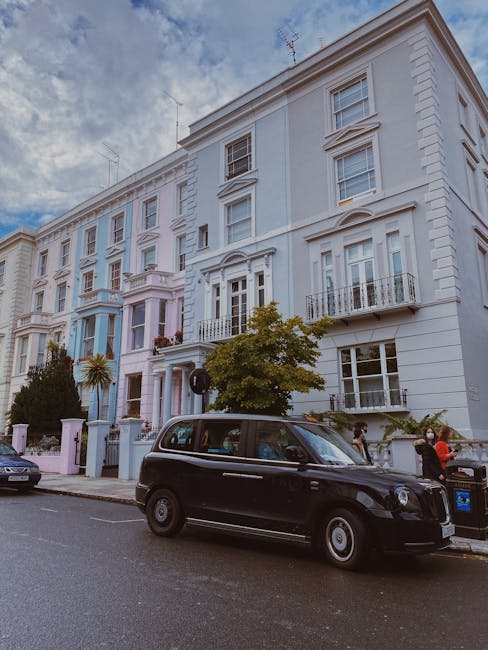 Image of a row of three-storey terraced houses with brightly painted façades in pastel pink, blue, and yellow, situated on a residential street with a pavement and metal railings. The houses have whi