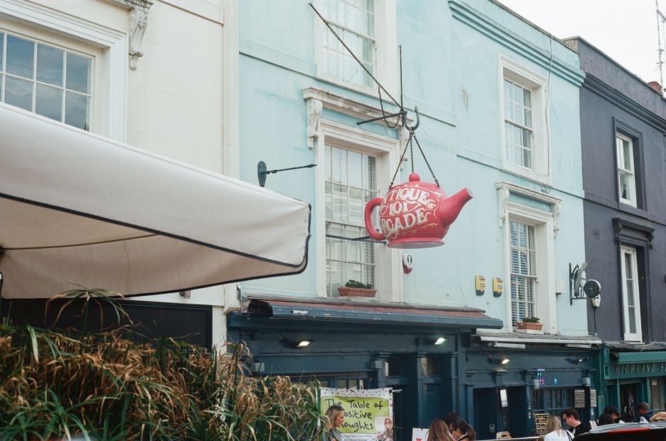 The image depicts the exterior of a building in a busy urban area, featuring a pastel blue facade with white window frames and decorative trim. A large, red teapot-shaped sign with white lettering han