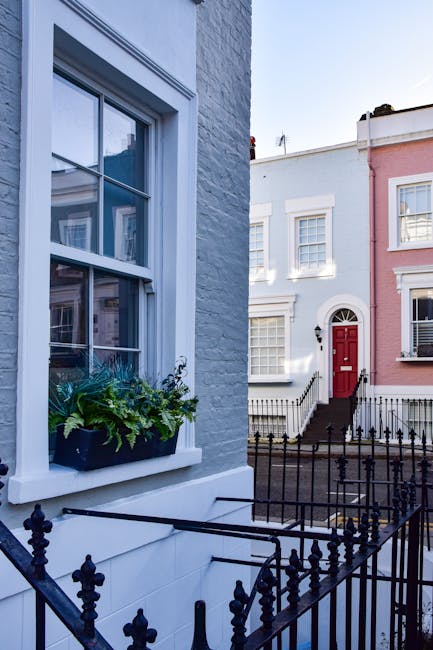 A white brick residential building with a large double-hung sash window featuring white trim and a window box filled with green plants and flowers. In the foreground, a black wrought iron railing surr