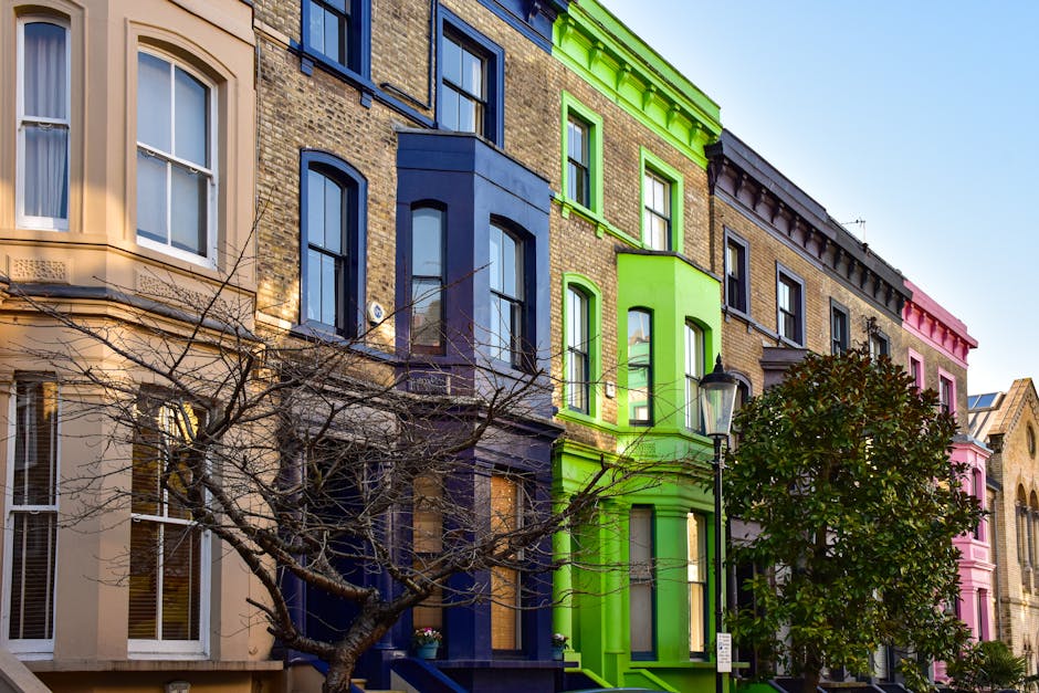 Exterior view of a row of residential terraced houses with varied facade colors and architectural details, including bay windows and decorative moldings, under a clear blue sky. In the foreground, a s