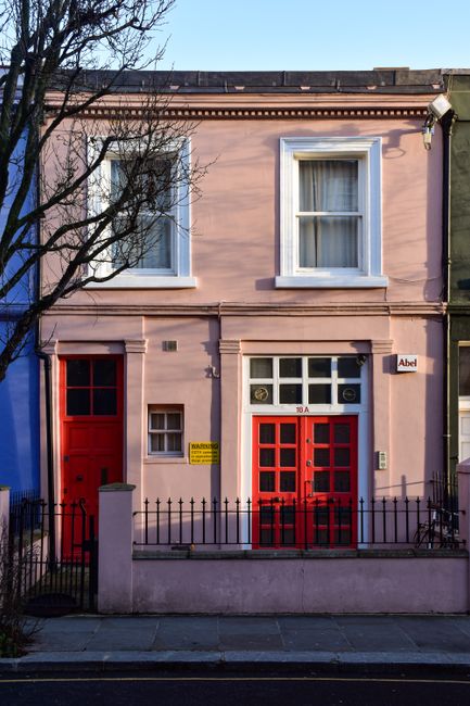 A two-storey townhouse with a pink facade, situated within a residential street. The building features large white-framed windows on the upper floor, and a smaller window on the ground floor next to a