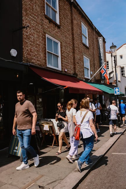 A busy street scene outside a three-storey brick building with large windows and a red and green striped awning, where pedestrians are walking along the pavement. Some individuals are carrying shopping bags while others are engaged in conversations or walking past. The building features a sign advertising antiques and collectibles, with an outdoor stand displaying framed artwork or posters nearby. A streetlamp and a flagpole with a Union Jack flag are visible on the corner, and the scene is illuminated by daylight, illustrating a typical urban environment suitable for home relocation or moving services. Self Storage Notting Hill may use such imagery to depict the vibrant community context for house removals or furniture transport.