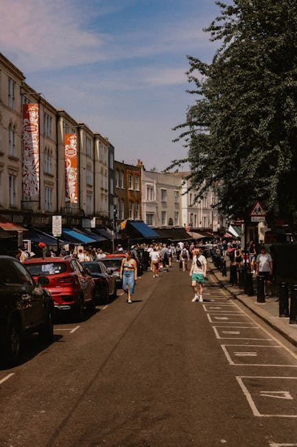 A busy street scene on Portobello Road in Notting Hill during a market day, with pedestrians walking along the pavement and browsing outdoor stalls covered by colorful umbrellas and canopies. The street is lined with Victorian-style terraced buildings featuring decorative facades and large windows, some displaying signs for shops and cafes. Union Jack flags and a banner reading 'Portobello Road W11' are strung across the street, adding a festive atmosphere. Several people are engaged in conversations or observing items, while others are moving briefly through the area. Vehicles are parked along the curb, and the overall scene is illuminated by natural daylight, with a partly cloudy sky overhead. In the context of house removals and home relocation, Self Storage Notting Hill might be supporting clients with packing or transportation, as evidenced by the busy street environment and the arrangement of goods typical of a relocation or moving process.