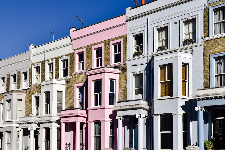 A row of Victorian-style terraced houses in Notting Hill, painted in pastel shades of teal, gray, pink, and white, with decorative columns and black wrought iron railings along the pavement. Some hous